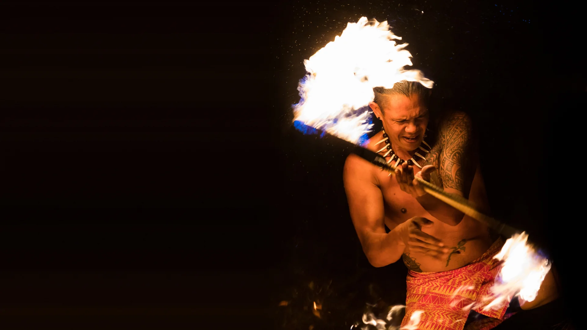 A fire knife dancer mid-spin at Luau Kalamaku, a blazing staff trailing flame against the night, wearing a tooth necklace and red patterned shorts