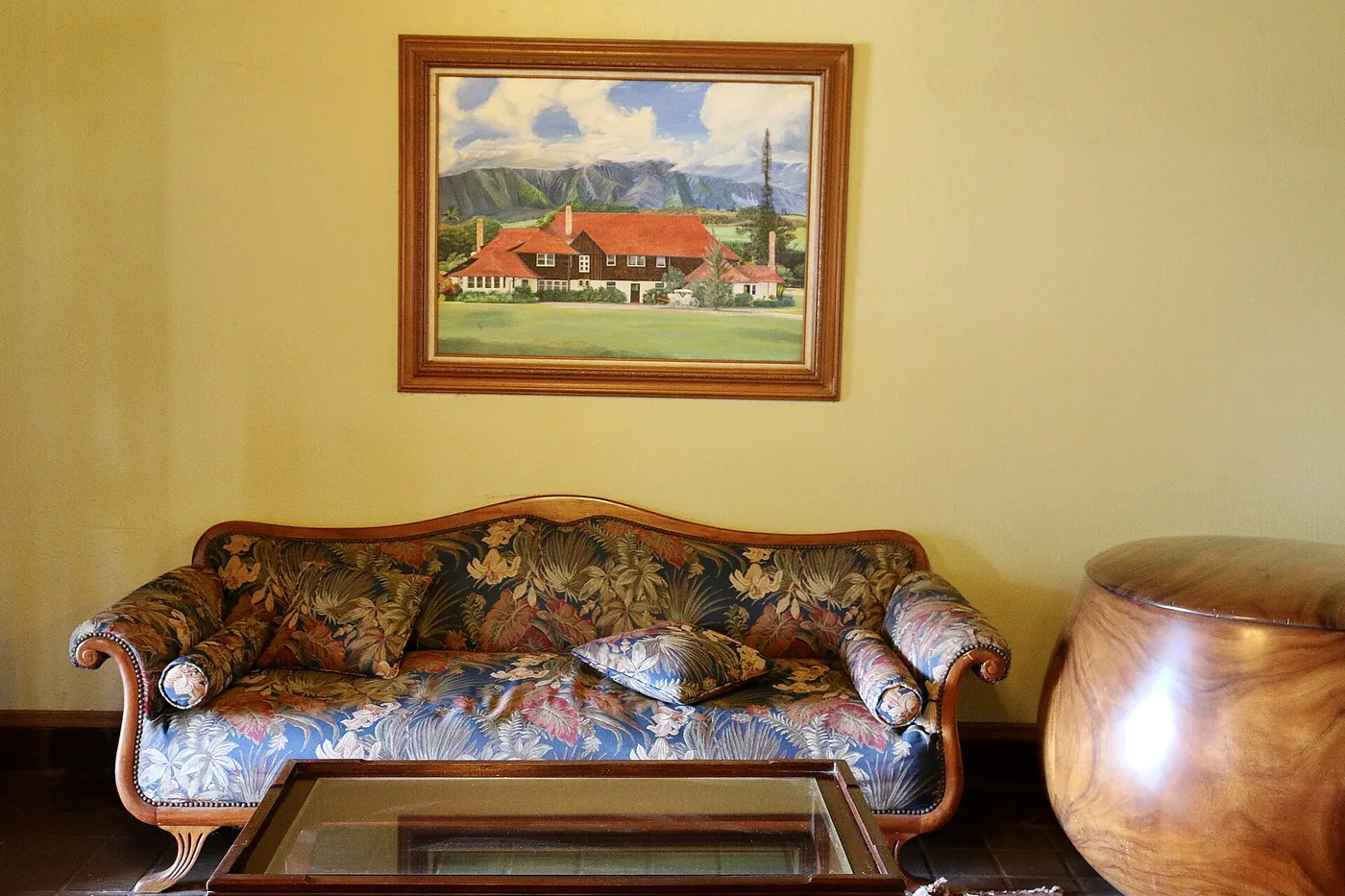 A floral-upholstered settee and polished wooden log drum beneath a framed painting of the manor house in a plantation parlour
