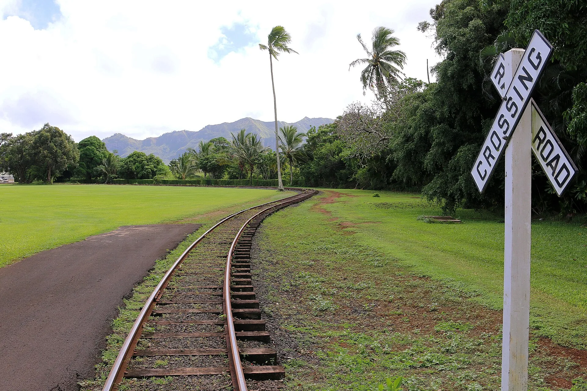 A curving plantation railway track through green lawn at Kilohana, with palms, a crossing sign and mountains in the distance