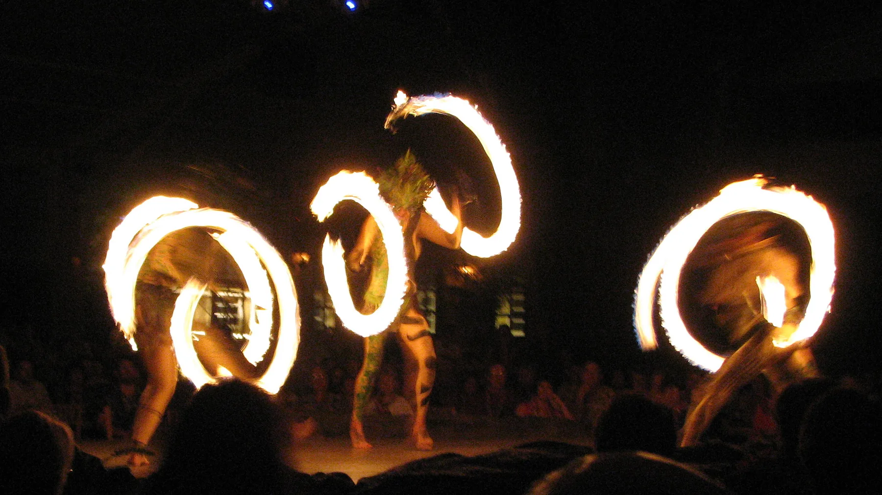 Three fire dancers on the Luau Kalamaku stage, each spinning a ring of flame in the dark while the audience watches